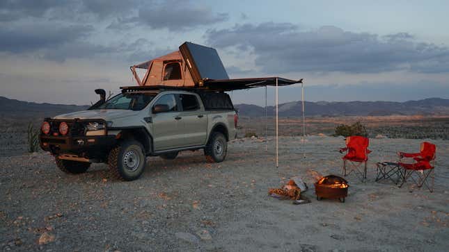A beige off-road truck with rooftop tent and awning is set up in the desert at sunset
